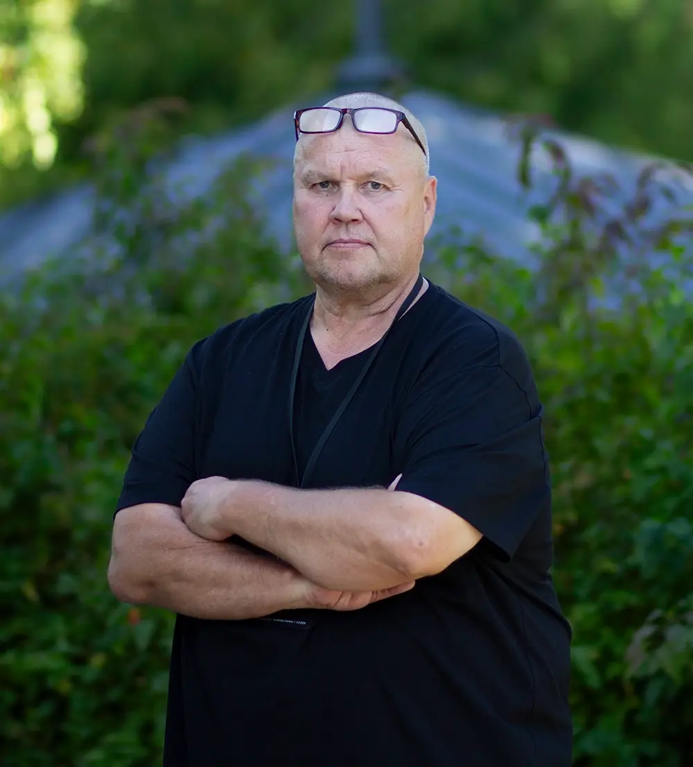 Timo Jutila katsoo vakavana kameraan kädet ristissä pensaiden edessä.