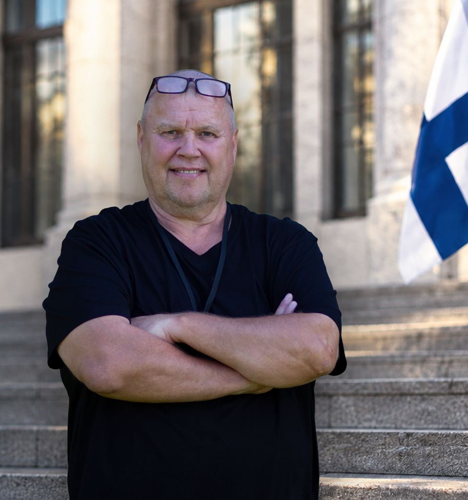 Timo Jutila seisoo kädet ristissä kivirakennuksen portailla. Taustalla liehuu Suomen lippu.
